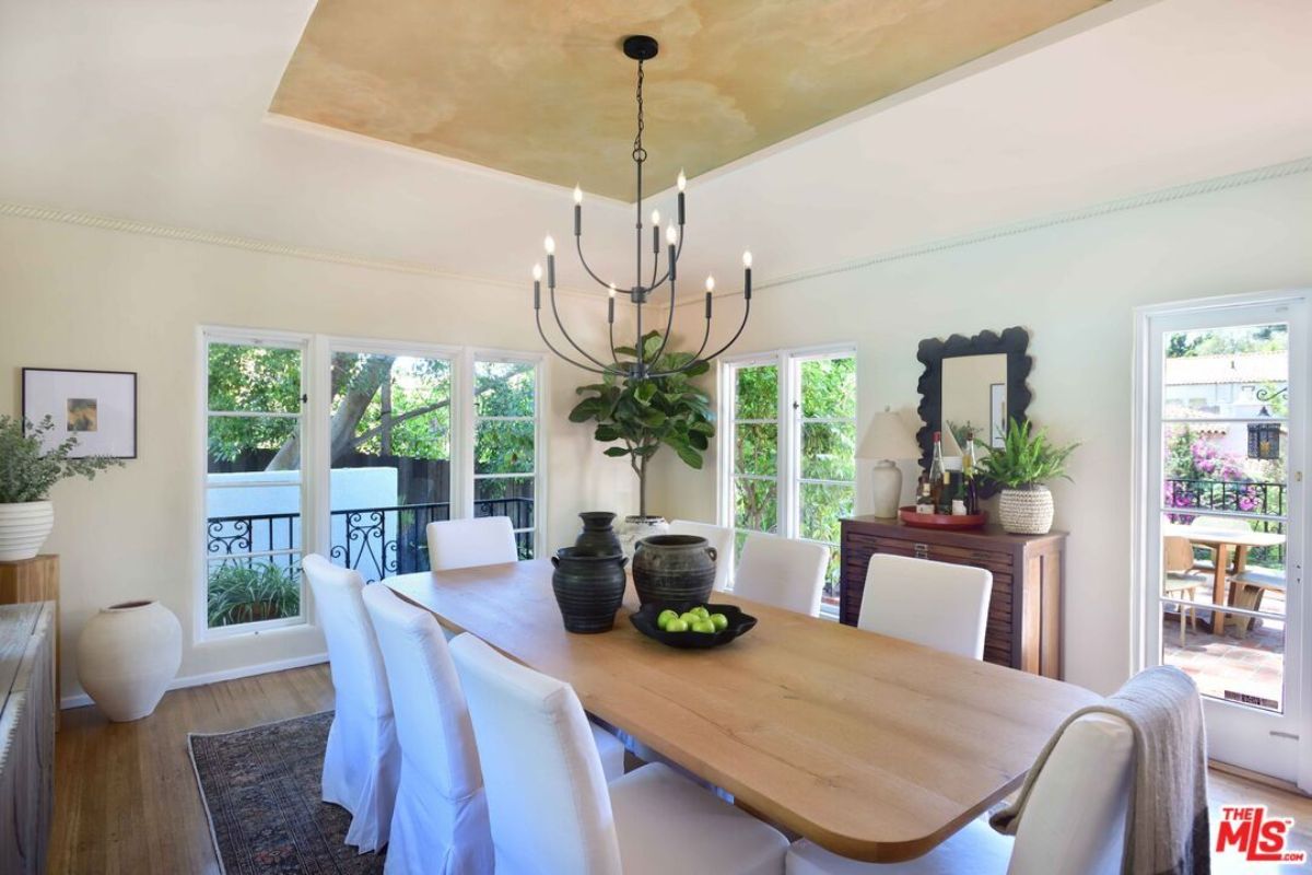 A dining room with houseplants, vases, windows, and a dining table surrounded by chairs.