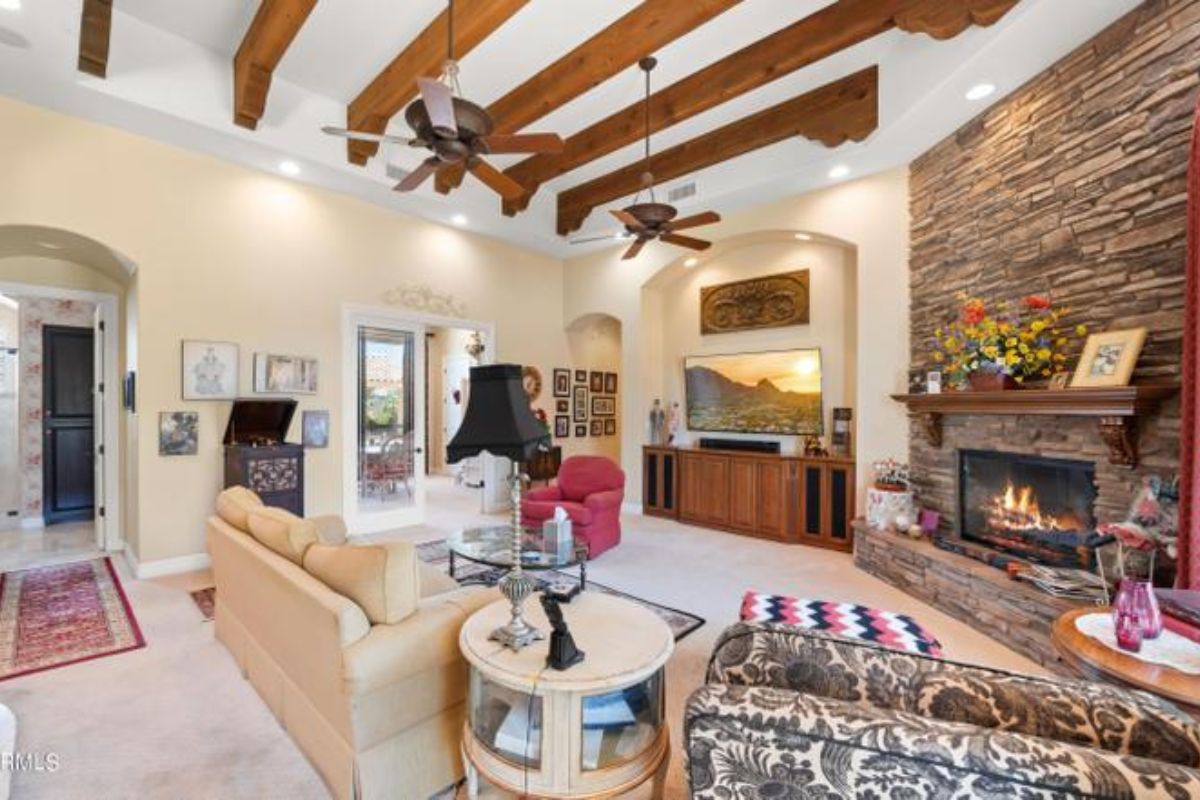 A living room with an exposed beam ceiling, ceiling fans, fireplace, cabinets, couches, and a glass center table.