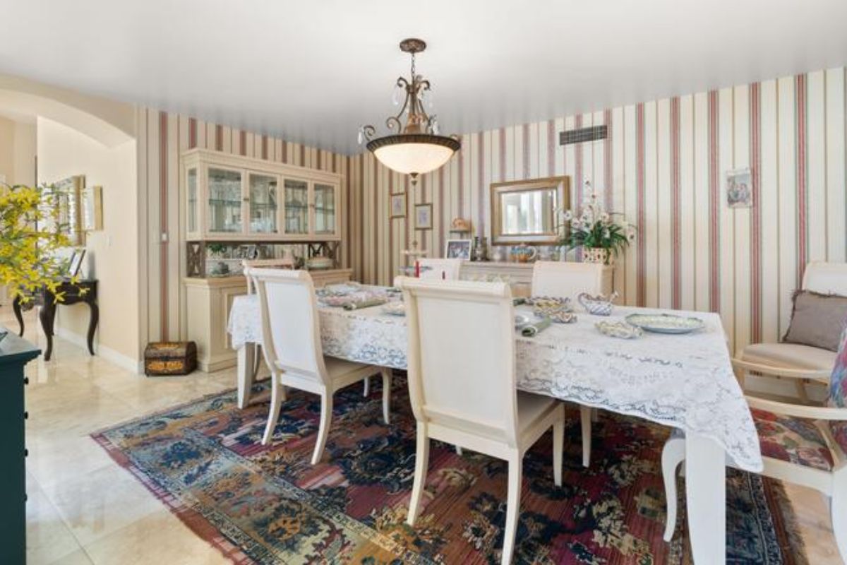 A dining room with wallpaper, houseplants, a rug, and a dining table surrounded by chairs.