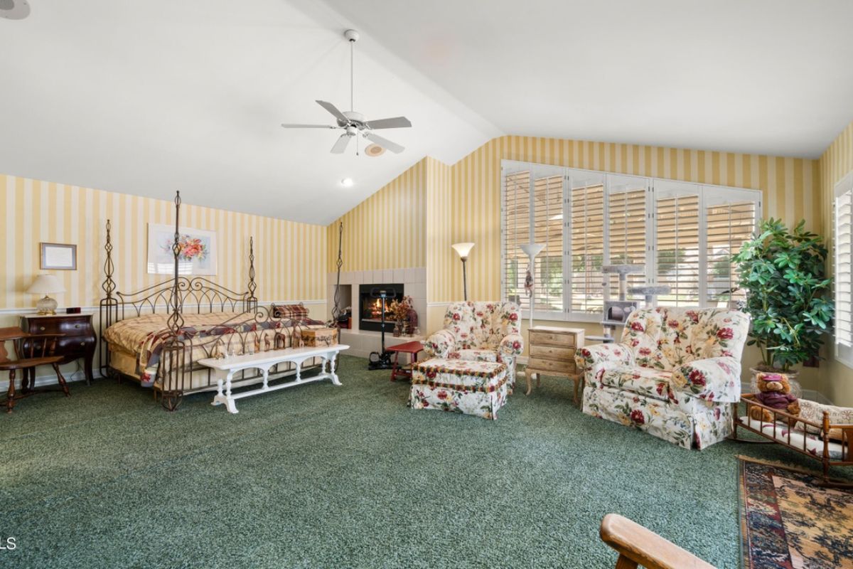 A bedroom with a bed, wallpaper, vaulted ceiling, ceiling fan, fireplace, chairs, and cabinets