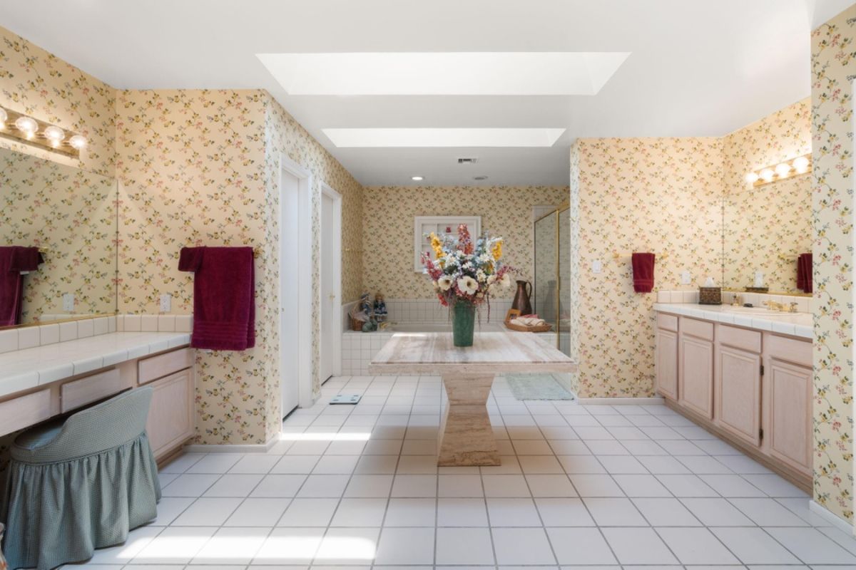 A bathroom with wallpaper, tiled floor, a center table, countertops, mirror, and cabinets