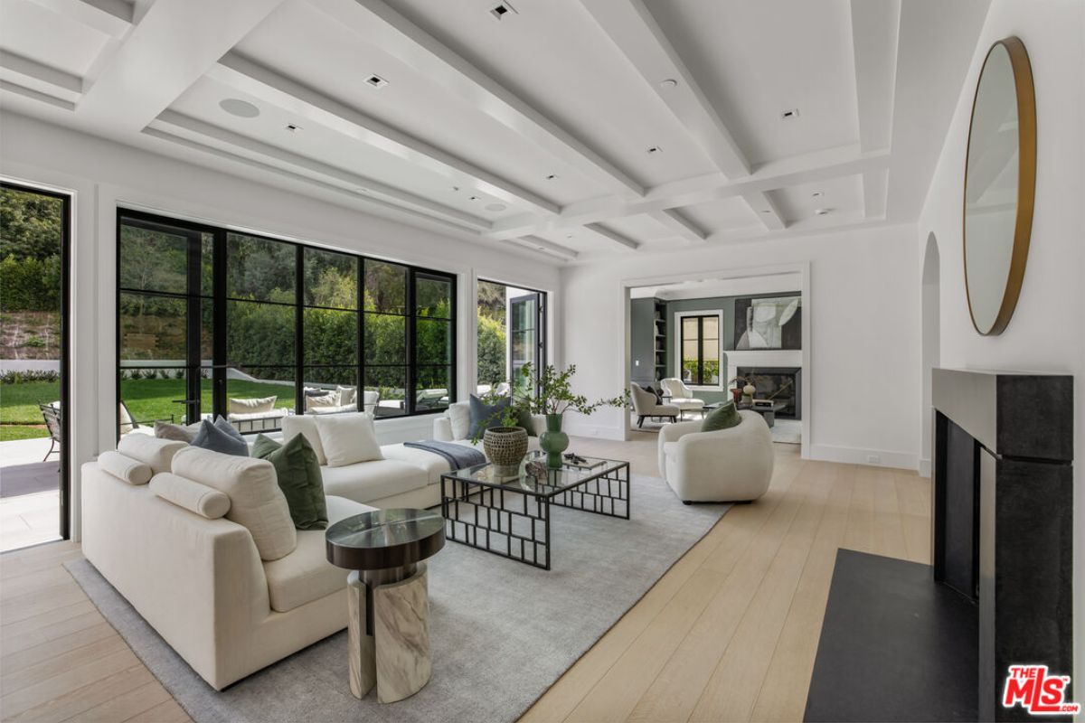 A living room with a fireplace, exposed beam ceiling, windows, white couches, and a glass center table.