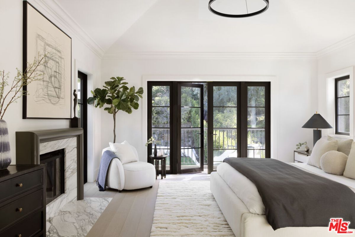 A bedroom with a bed, fireplace, painting, cabinet, chair, and houseplants.