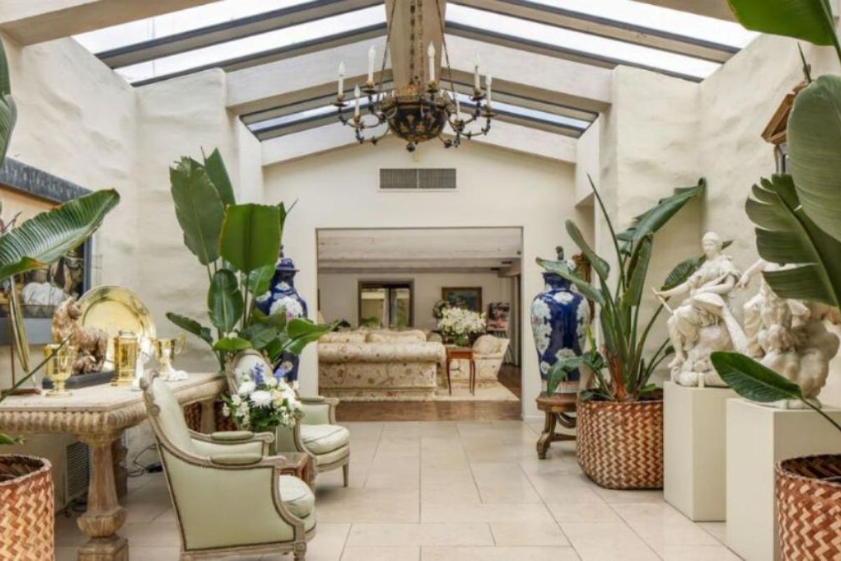 A sunroom with skylight roofing, a chandelier, houseplants, statues, a table, chairs, and paintings.