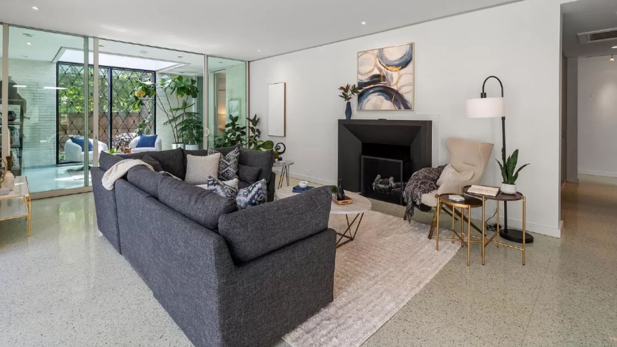 A living room with a rug, couch with pillows, houseplants, coffee tables, painting, and fireplace.