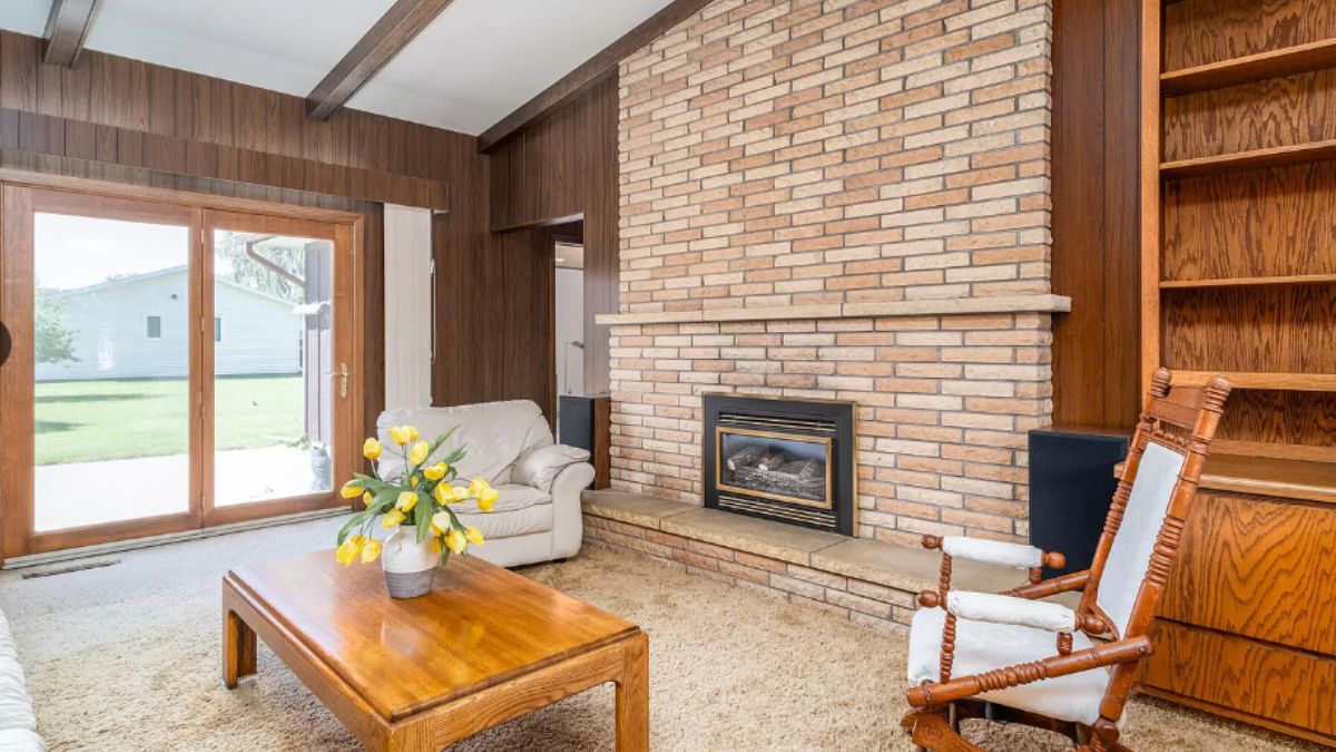 A family room with a brick fireplace, cabinet, shelf, studio couch, chair, and a center table.