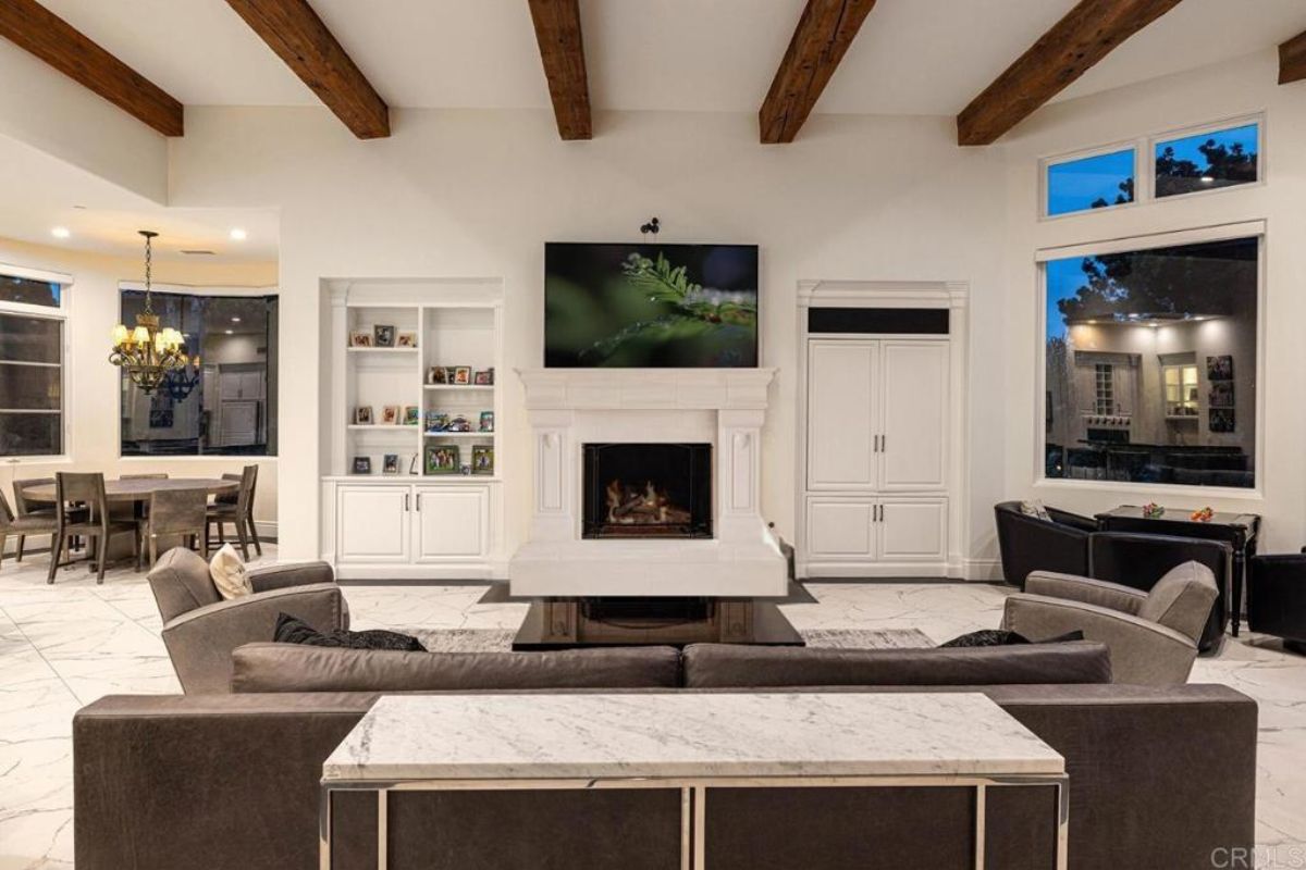 A living room with an exposed beam ceiling, fireplace, TV, shelf, and cabinets.