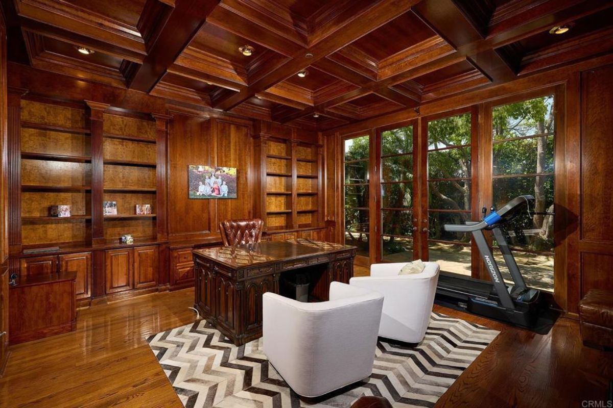 A home office with an exposed beam ceiling, shelves, fireplace, TV, studio couch, treadmill, chairs, and desk.