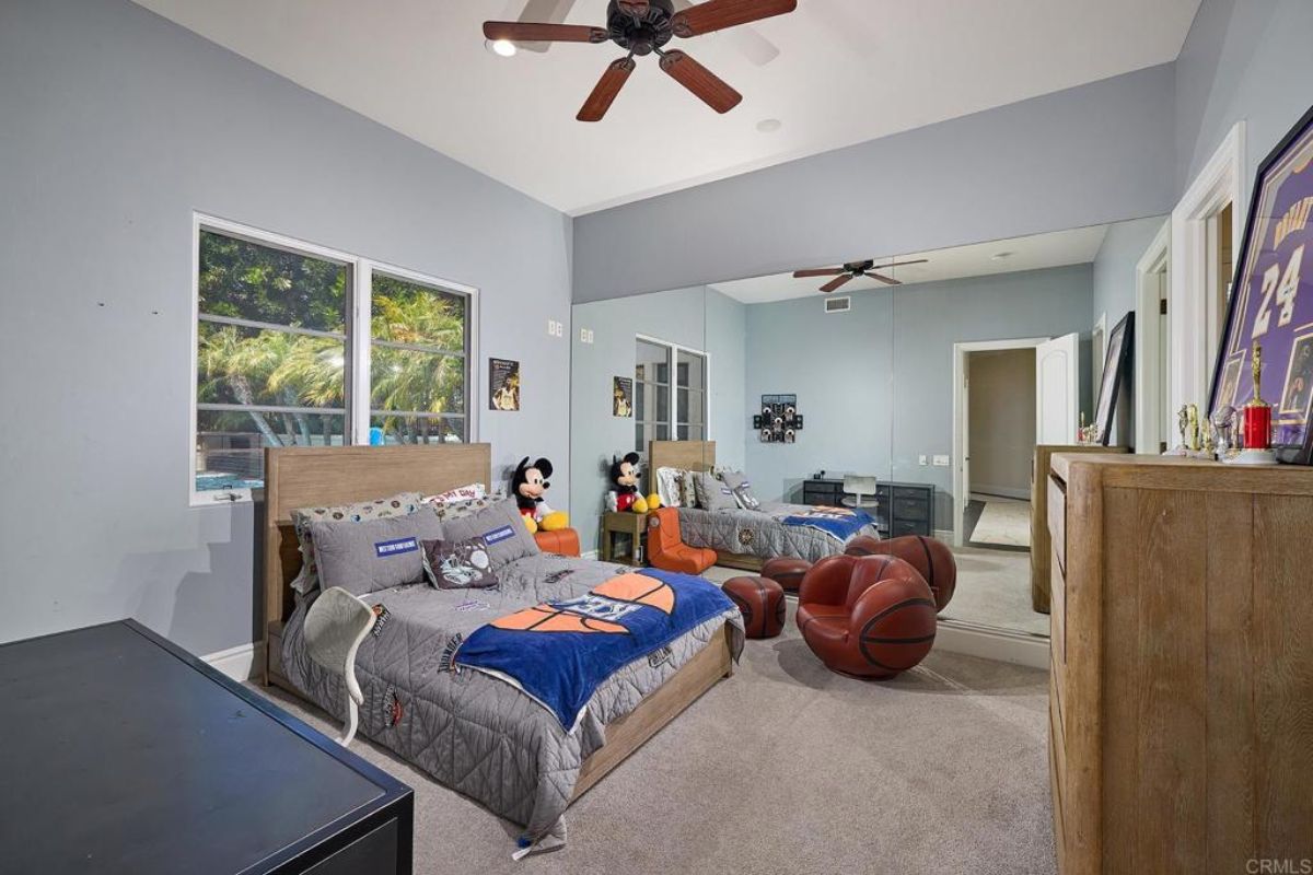 A bedroom with a bed, window, ceiling fan, chairs, cabinet, and mirror.