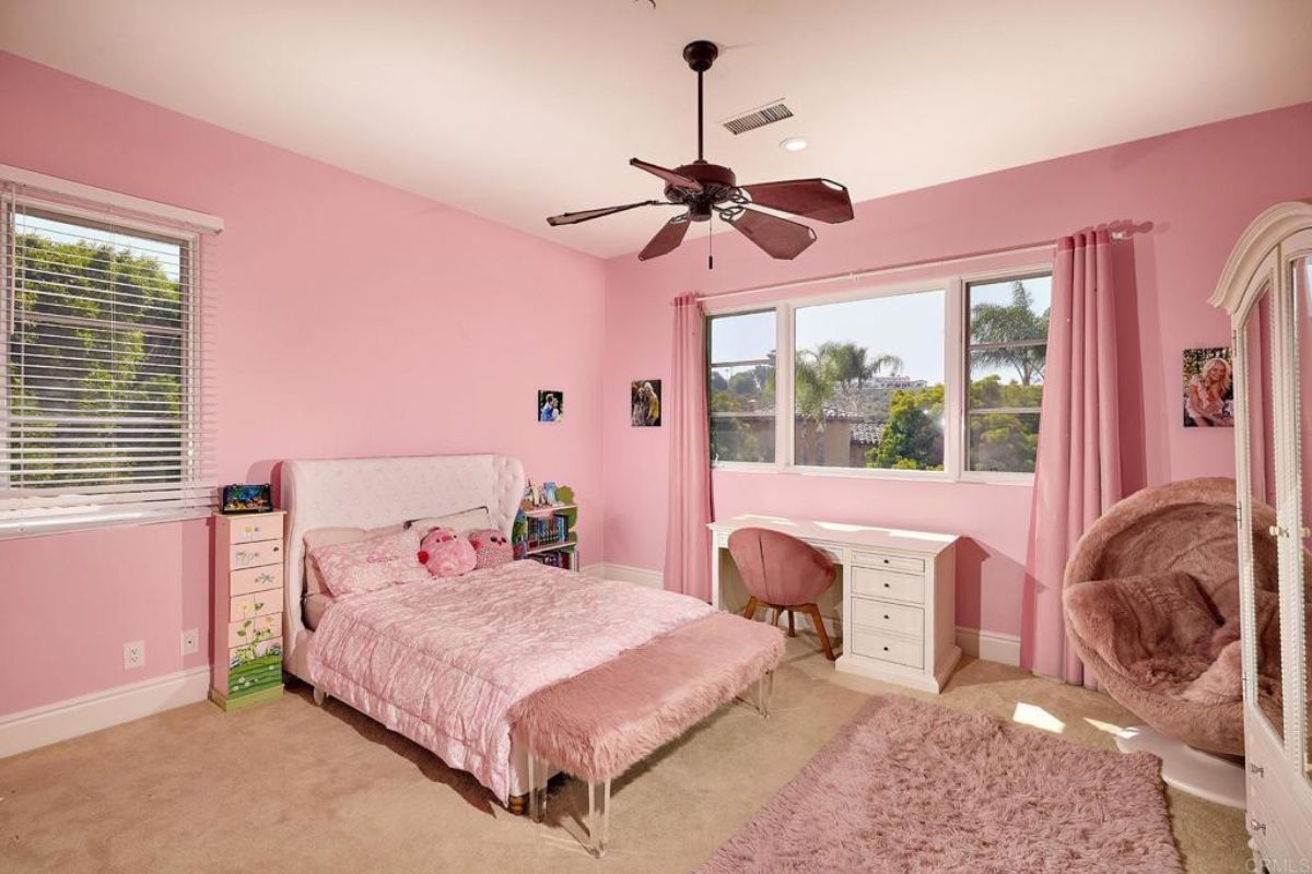 A bedroom with a bed, windows, a ceiling fan, pink walls, a desk, and a chair.