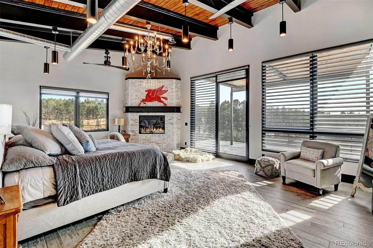 Primary bedroom with an upholstered bed and a corner fireplace under a beamed ceiling with exposed pipes.