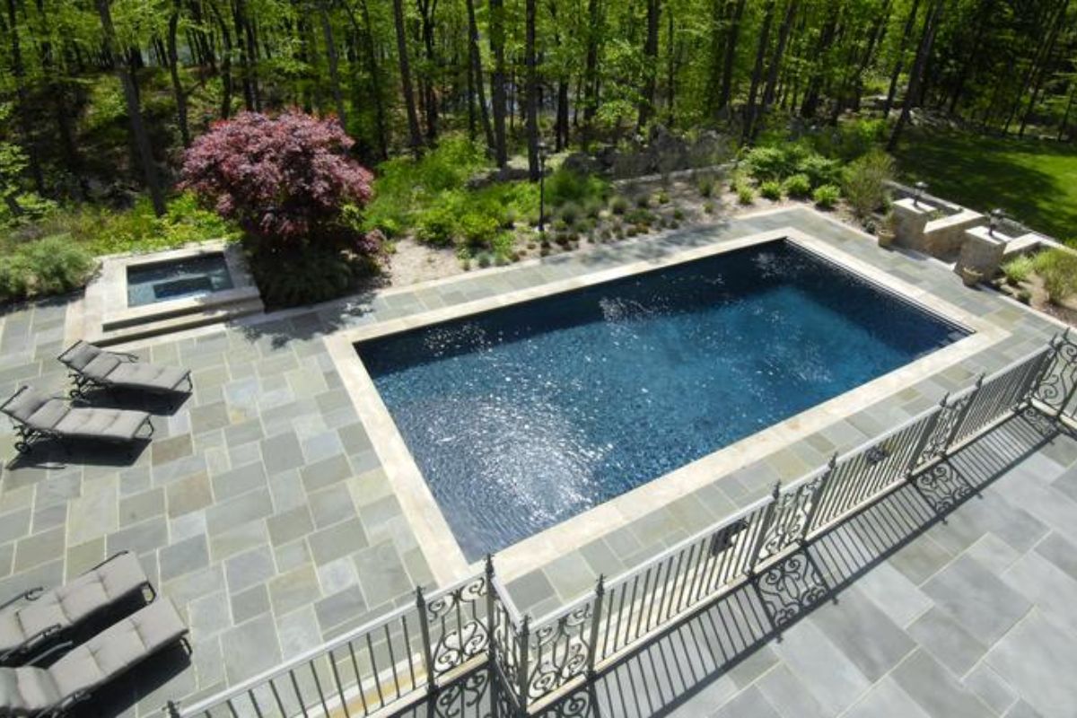 A swimming pool with a jacuzzi and chairs on the poolside, surrounded by trees.