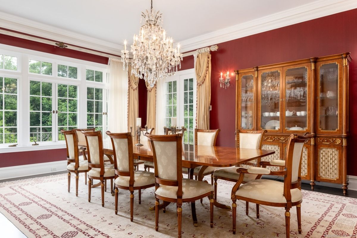 A dining room with windows, a chandelier, a rug, a cabinet, and a dining table surrounded by chairs.