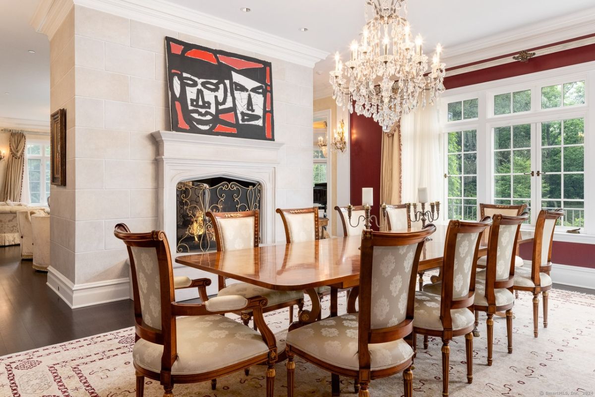 A dining room with windows, a chandelier, a rug, a fireplace, and a dining table surrounded by chairs.