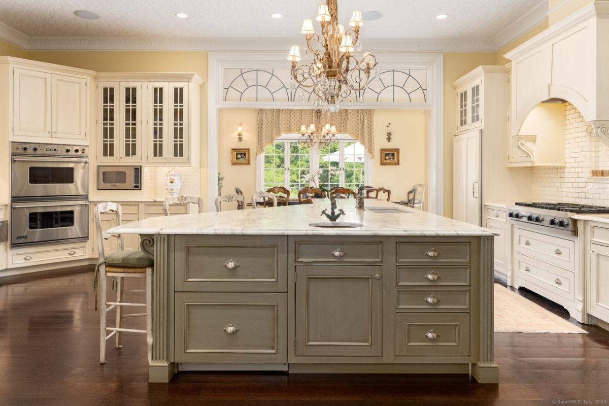 A kitchen with a chandelier, countertops, chairs, sink, kitchen appliances and cabinets.