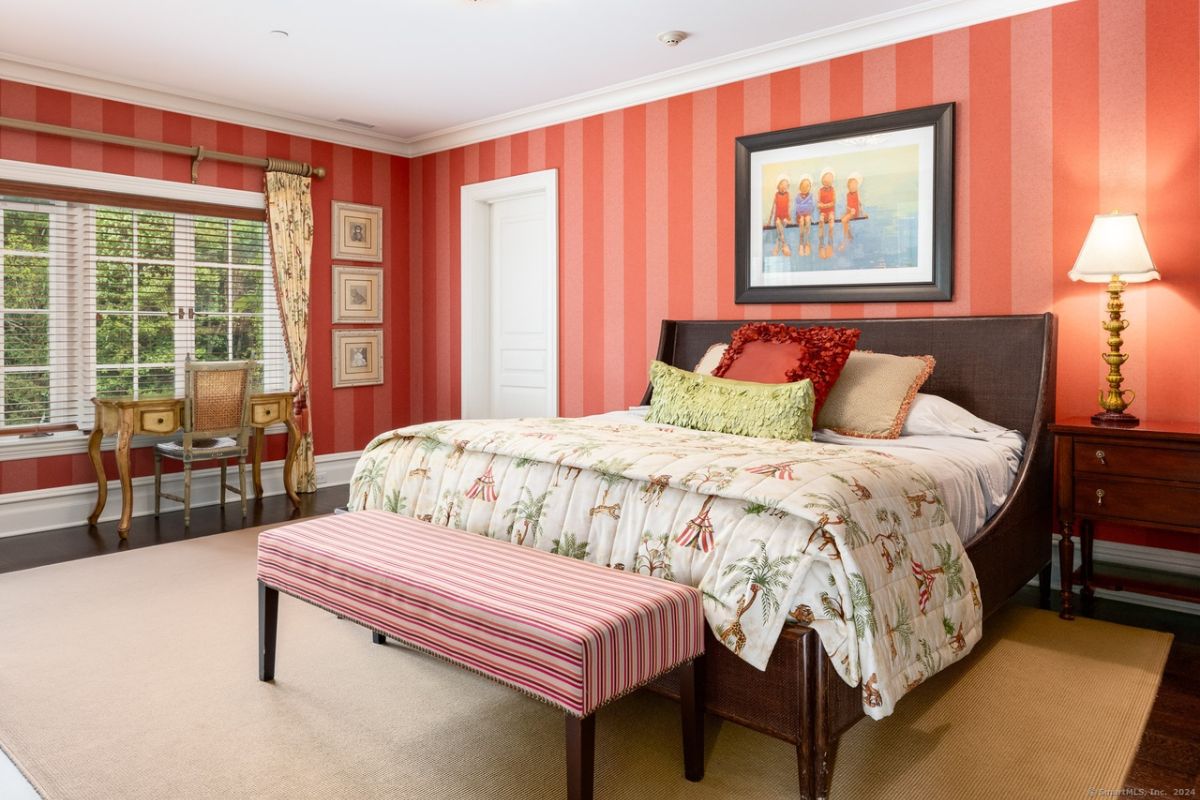 A bedroom with a bed, wallpaper, a rug, a nightstand, a desk, and a chair.