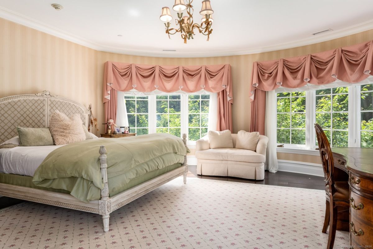 A bedroom with a bed, chandelier, windows, a couch, desk, and chair.