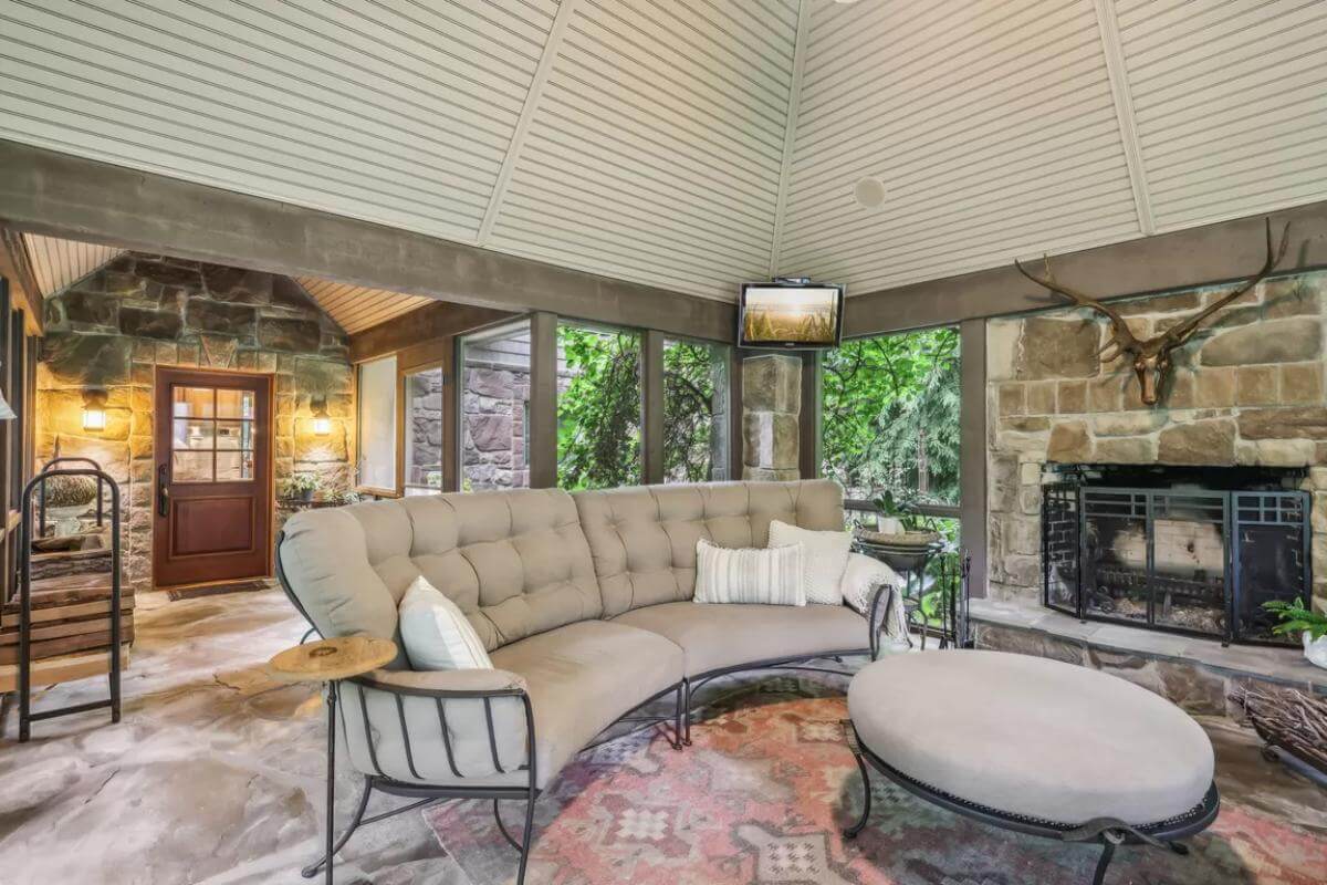 Covered porch with a tufted sofa, a matching ottoman, and a stone fireplace.