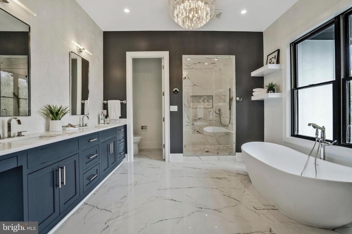 A bathroom with tiled floor, dual vanity, mirrors, cabinets, chandelier, and bathtub.