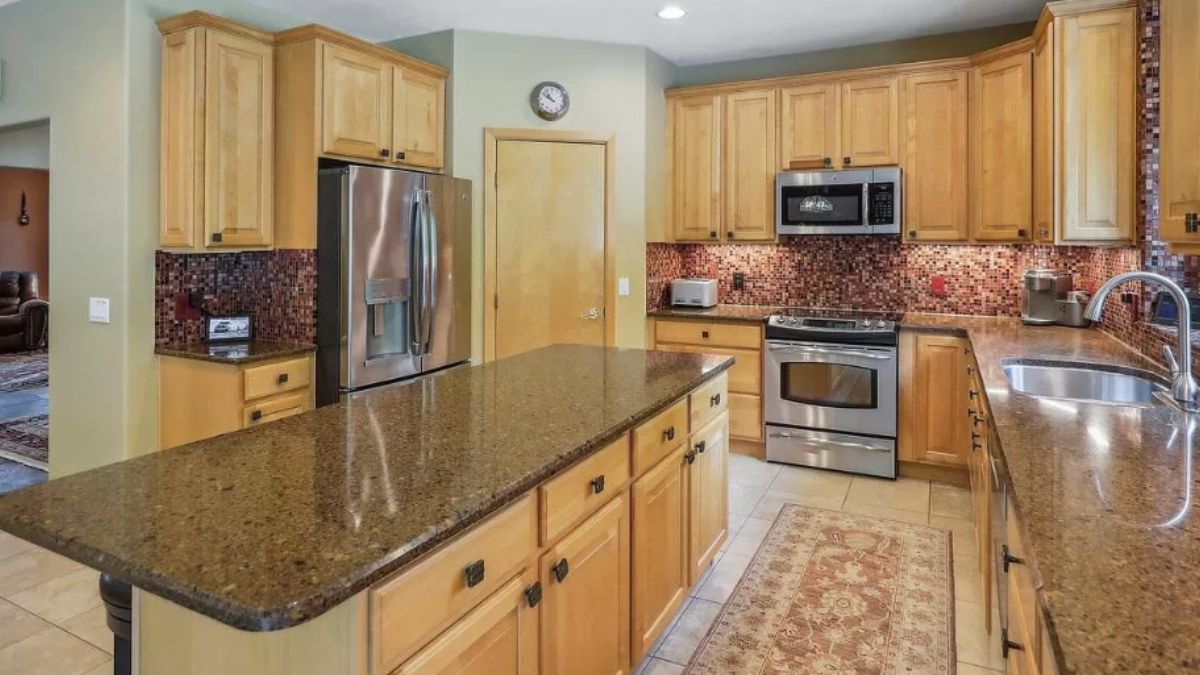 A kitchen with an island, stools, rug, countertop, sink, stove, and cabinets.