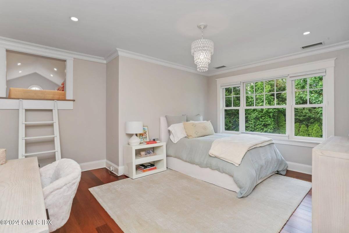 Bedroom with white furnishings and a trio of windows overlooking the lush yard.