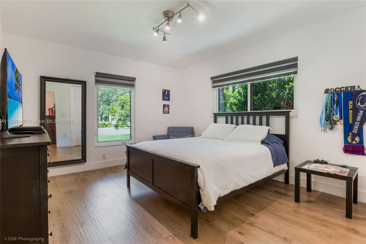 A bedroom with a bed, a TV, a cabinet, a side table, windows, and a mirror.