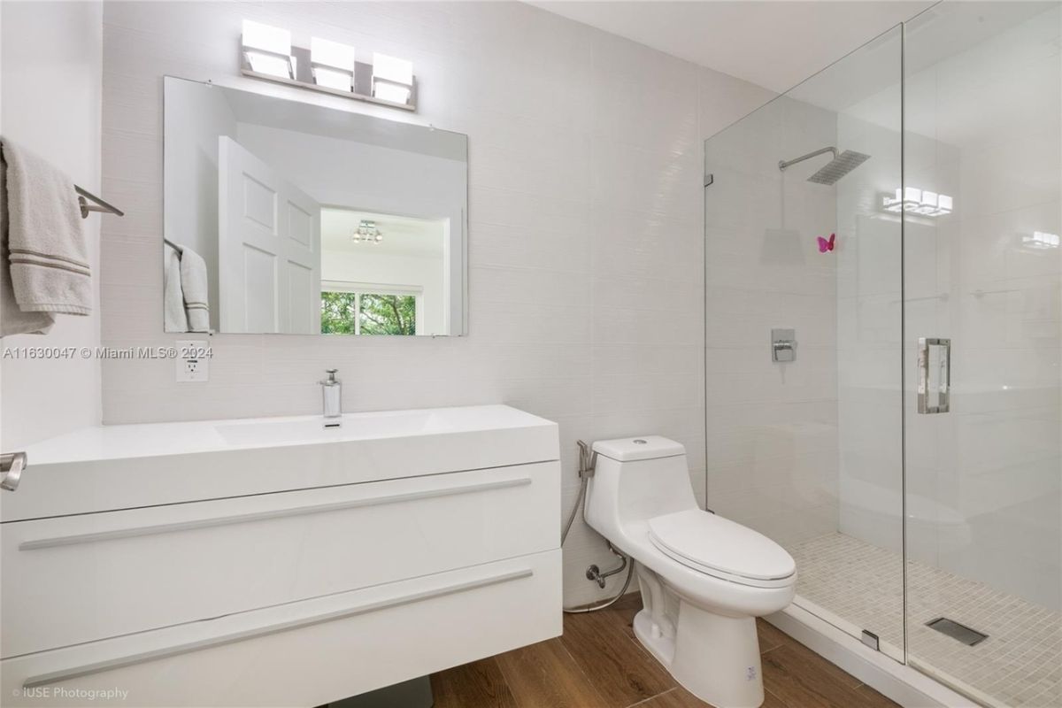 A bathroom with a vanity, a mirror, a toilet, and a shower room.