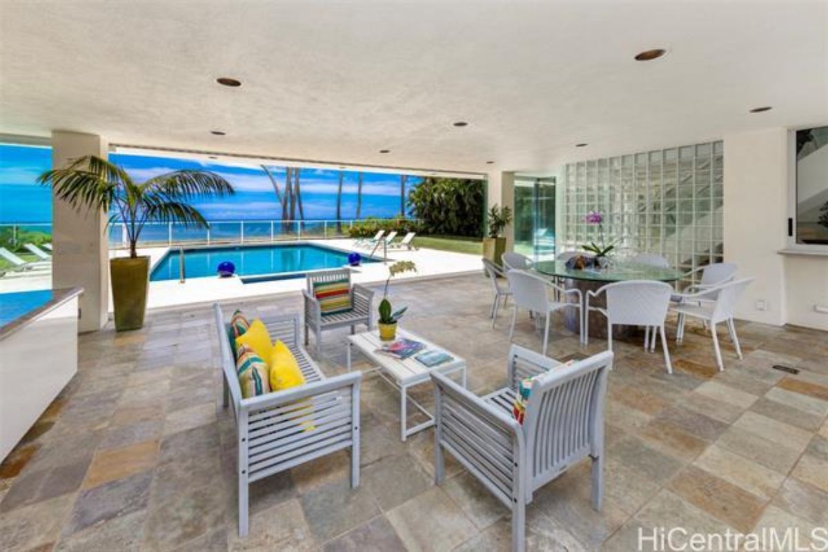 An outdoor living area with a bench, chairs, center table, and a glass dining table surrounded by chairs.