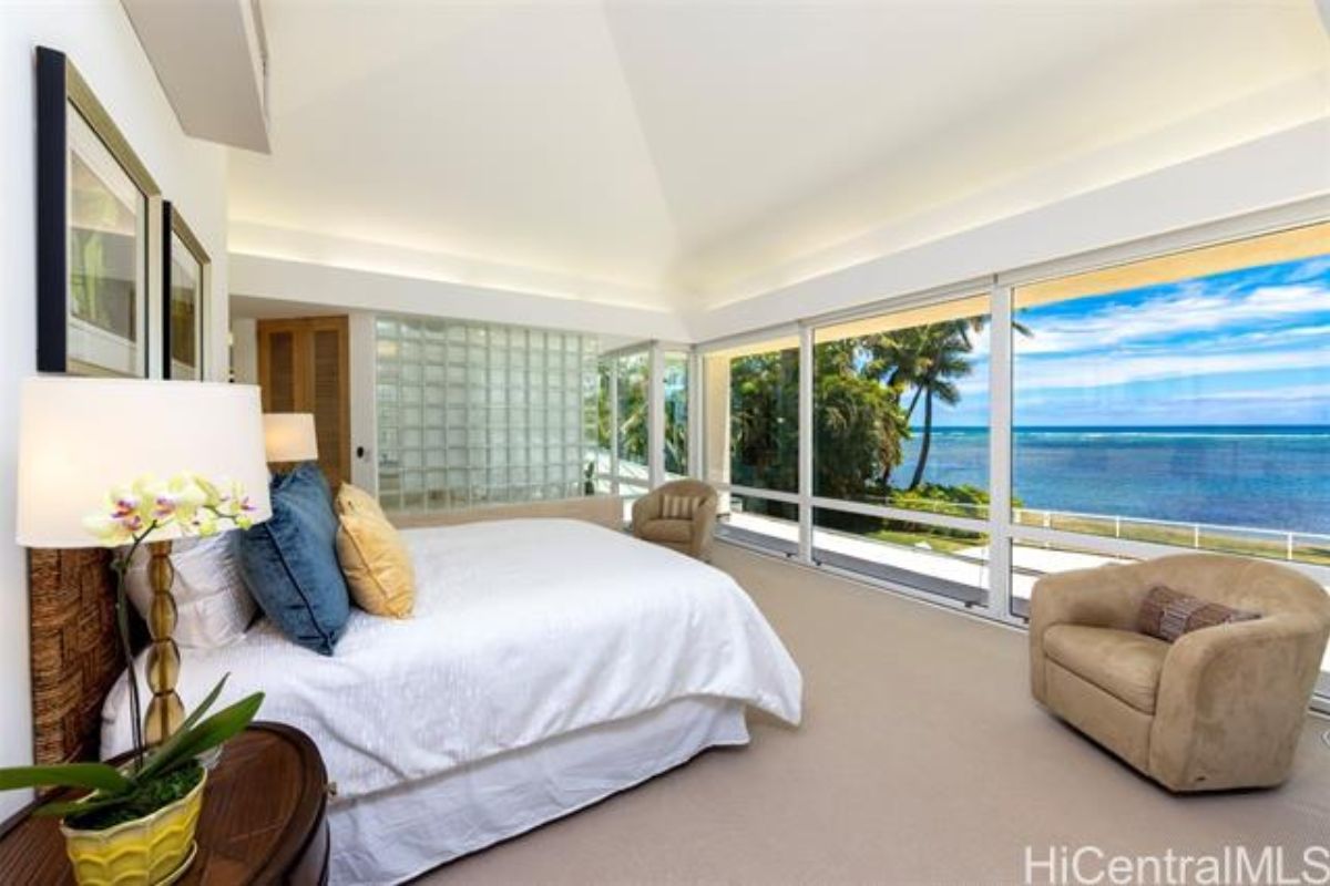 A bedroom with floor-to-ceiling windows, chairs, a nightstand, and a bed.