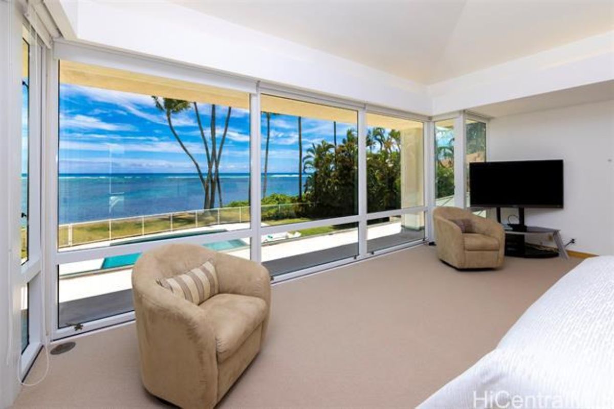 A bedroom with floor-to-ceiling windows, chairs, a TV, and a bed.