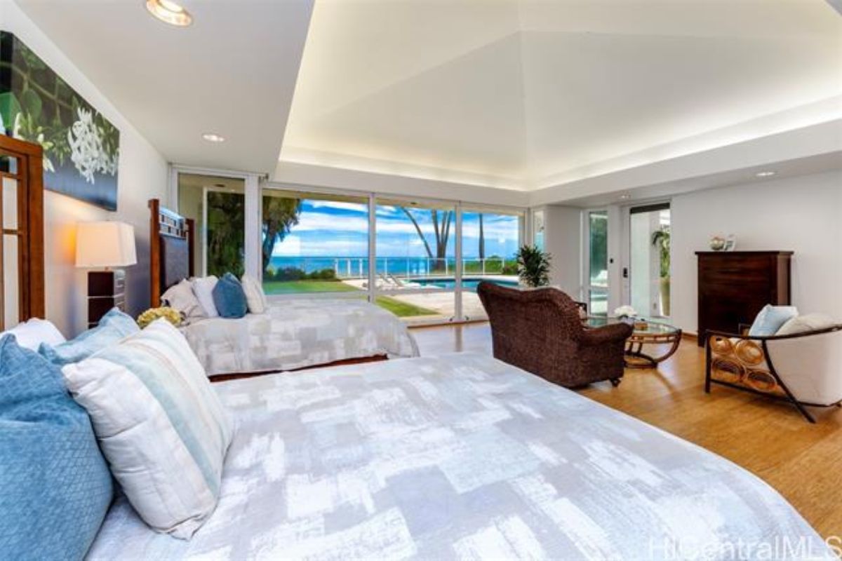 A bedroom with two beds, floor-to-ceiling windows, chairs, a nightstand, a cabinet, and a center table.