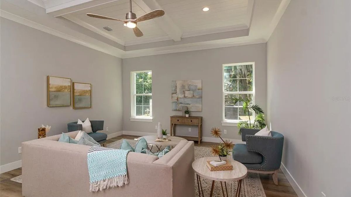 A living room with an exposed beam ceiling, ceiling fan, windows, paintings, couch, chairs, and coffee tables.