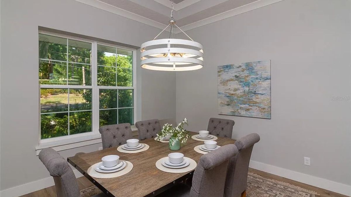 A dining room with a painting, window, chandelier, and a dining table surrounded by chairs.