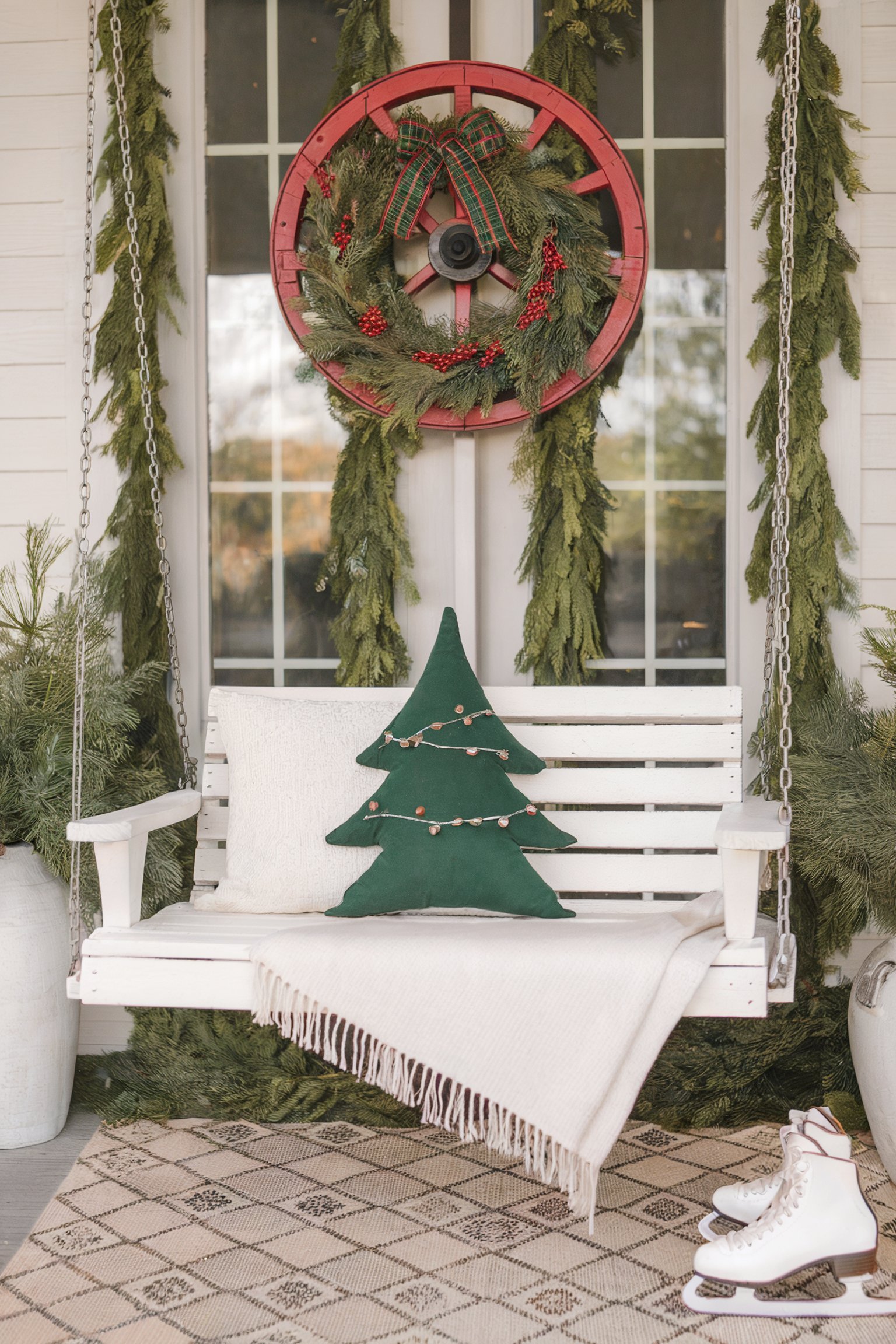 Porch swing with a Christmas tree pillow, a wagon wheel wreath, garland, and ice skates on a rug below