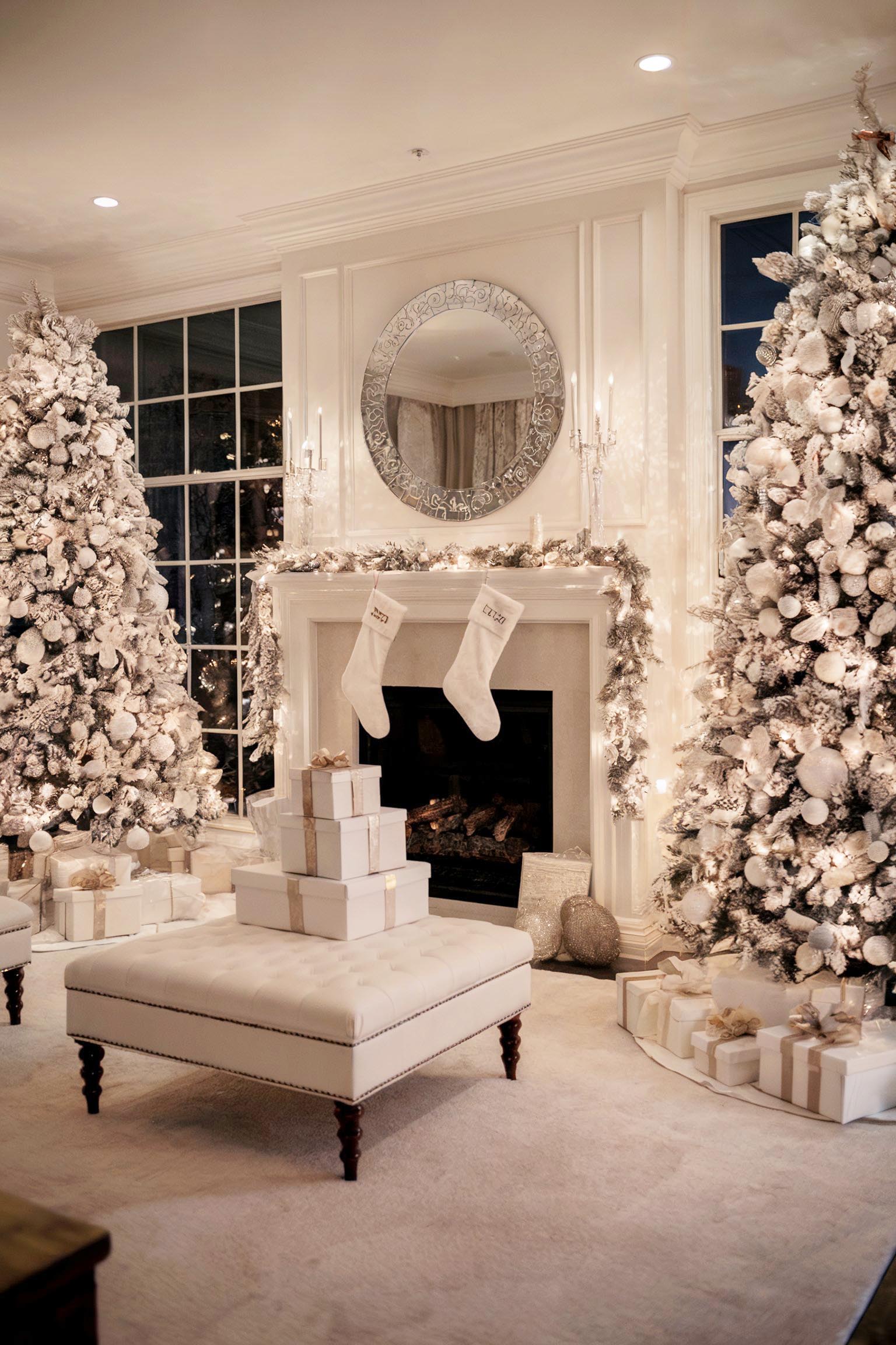 Two flocked trees, a fireplace with white stockings, garland, and a tufted ottoman topped with wrapped gifts