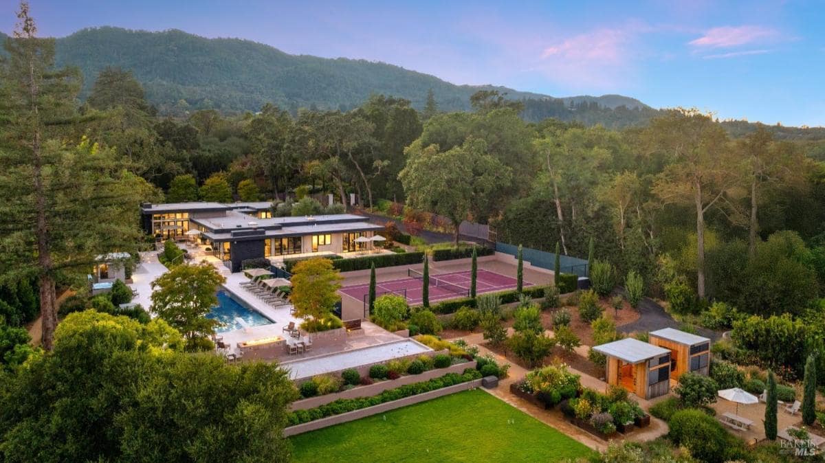 A modern home with a swimming pool, tennis court, landscaped gardens, and surrounding trees, set against a mountain backdrop.