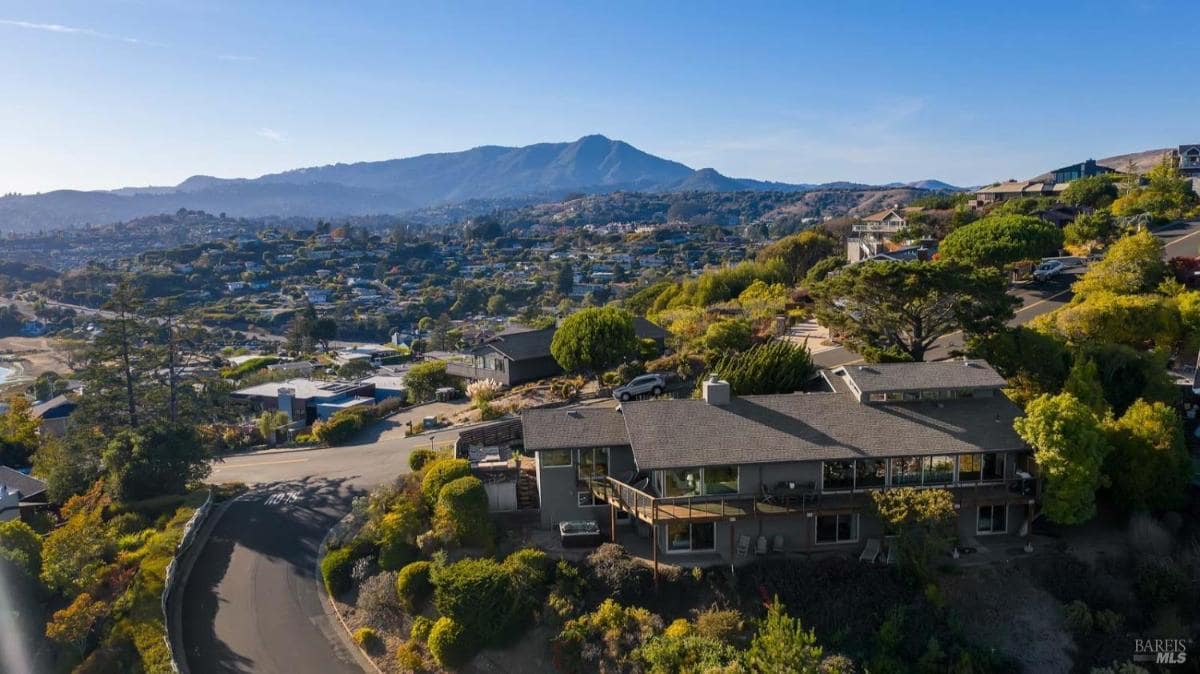 An aerial view of a hillside neighborhood with a large home, surrounding greenery, and distant mountains.