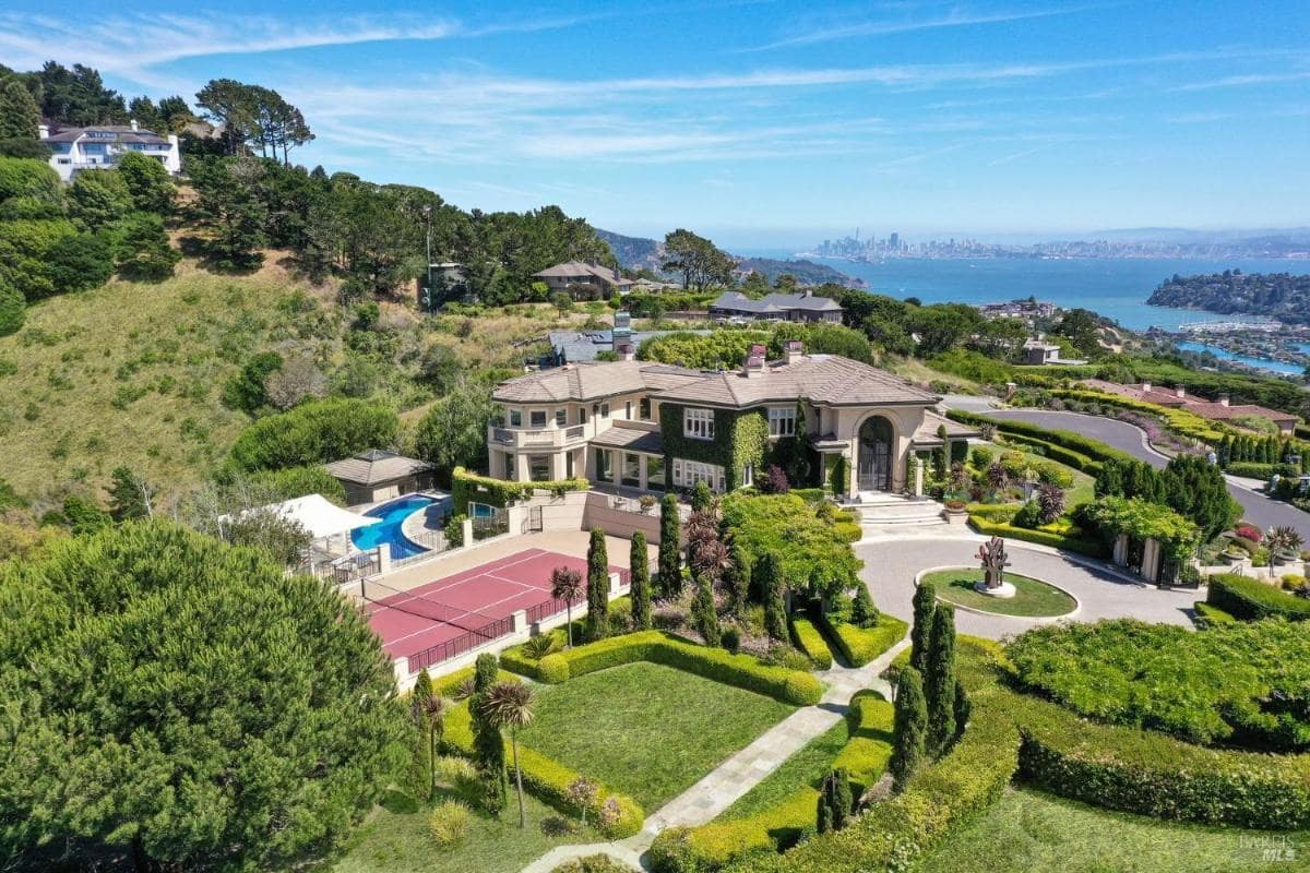 Aerial view of a luxury mansion surrounded by lush greenery, featuring a swimming pool, tennis court, and landscaped gardens.