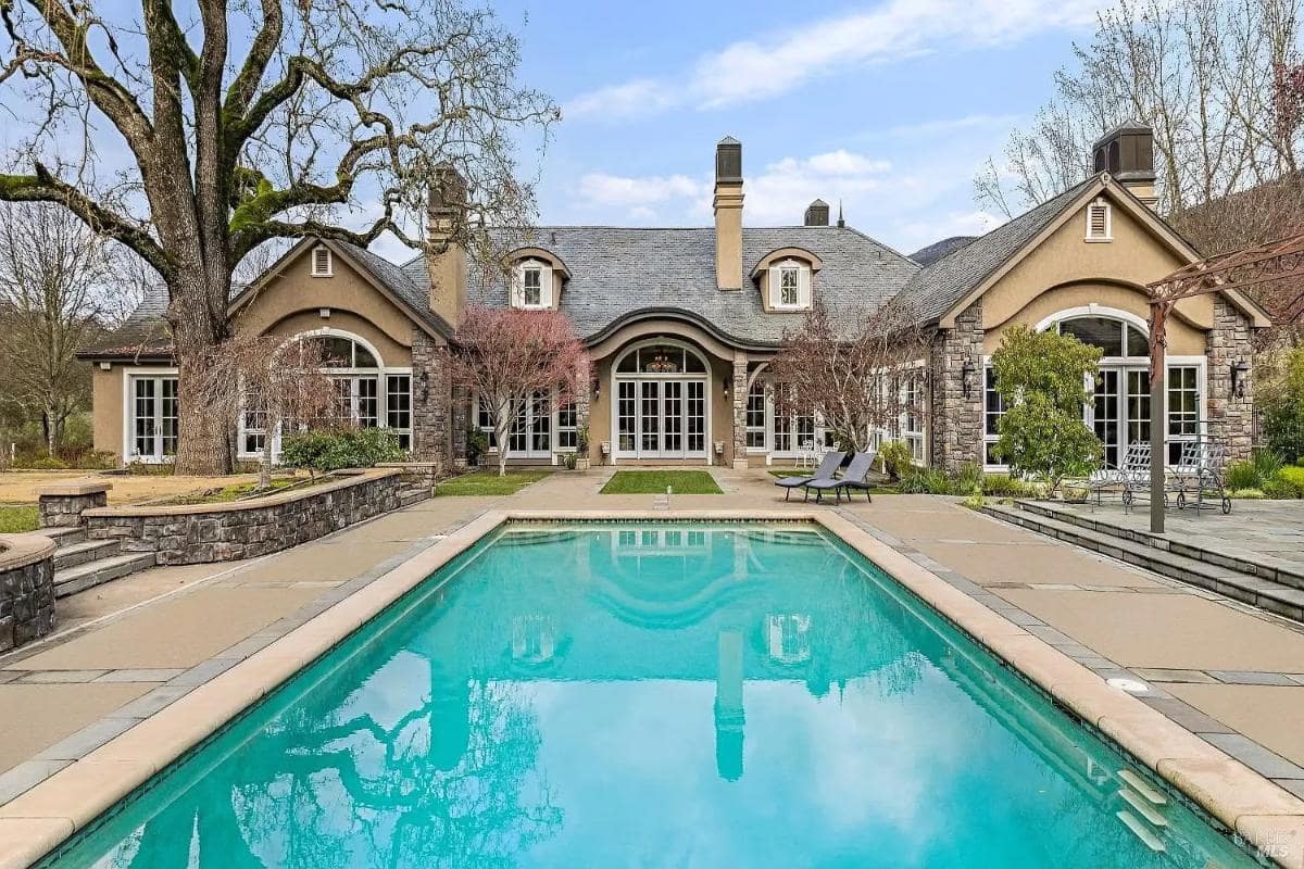 A large mansion with a stone facade, multiple arched windows, and a swimming pool in the foreground.