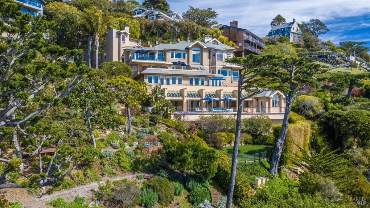 Multi-level hillside home with large windows, multiple balconies, and surrounding lush landscaping.