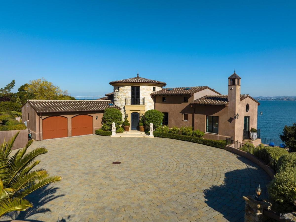 Mediterranean-style home with a circular entry, red garage doors, and a cobblestone driveway overlooking the waterfront.