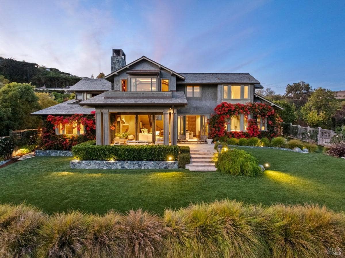 Two-story gray house with large windows, front porch, and red flowers. Landscaped yard with grass and shrubs.