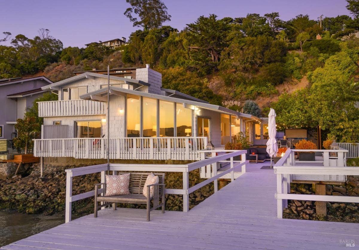 Exterior of a waterfront home with a large deck and hillside backdrop.