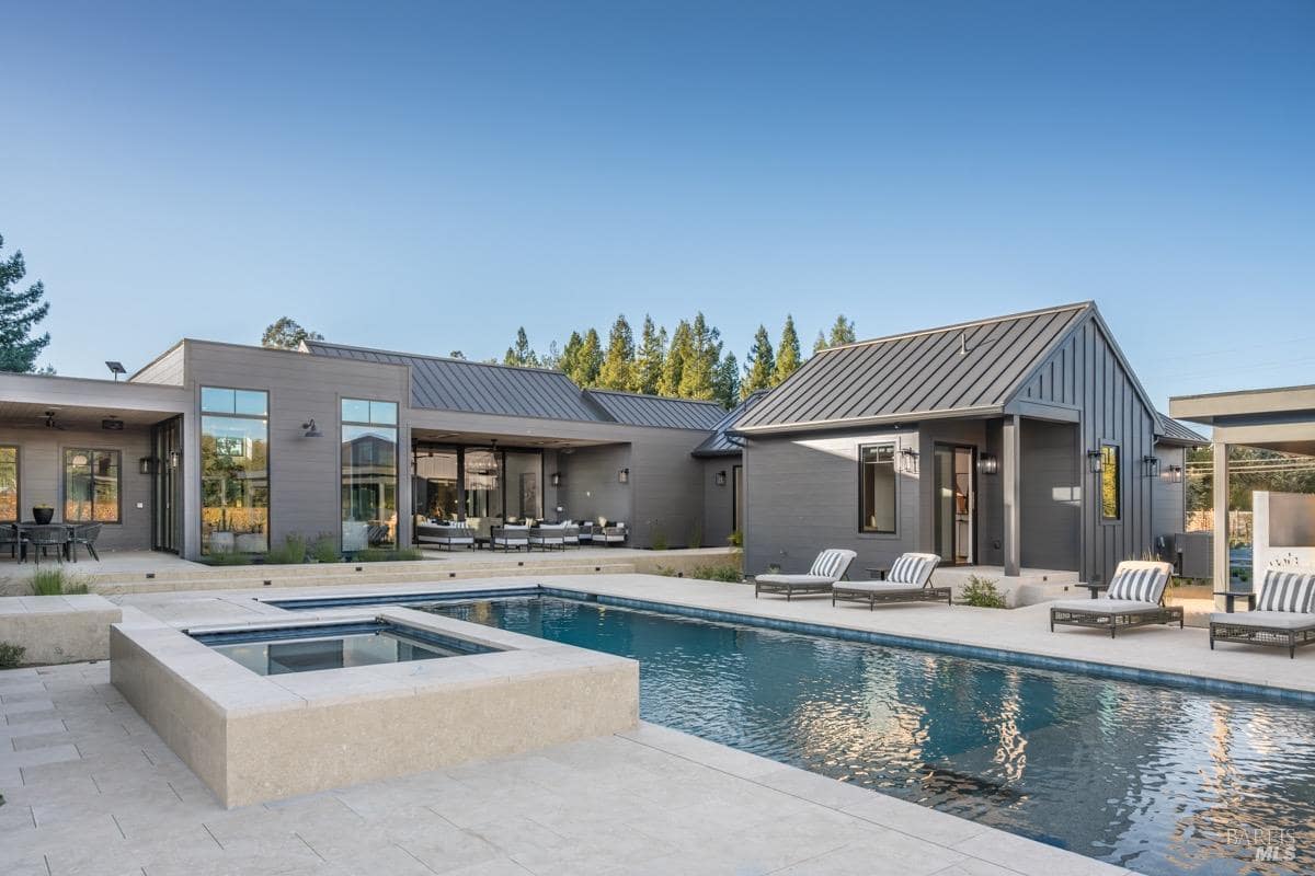 A modern home with gray exterior siding, metal roof, and large pool area featuring lounge chairs, a spa section, and expansive patio space.