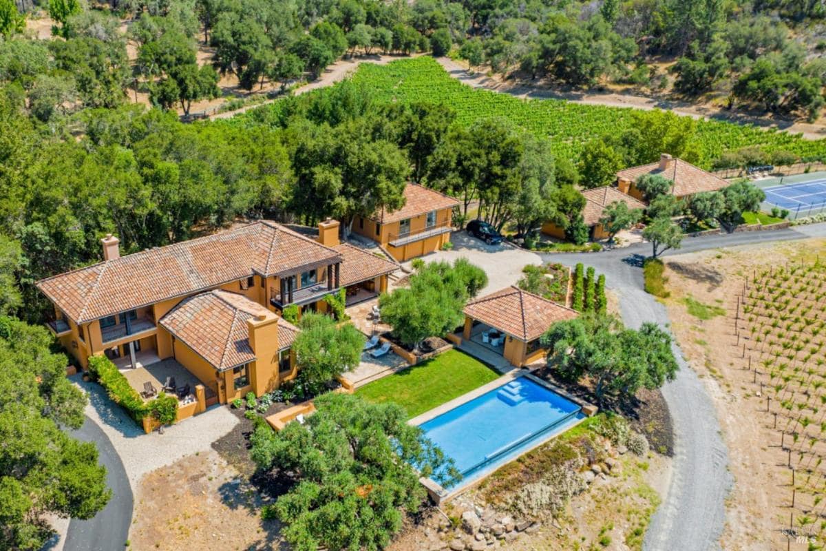 Aerial view of a house with multiple buildings, a swimming pool, and surrounding vineyards.