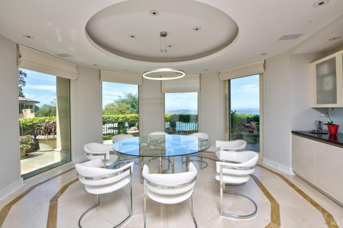 A modern circular breakfast nook with a glass table, white chairs, and expansive windows showcasing the surrounding garden.