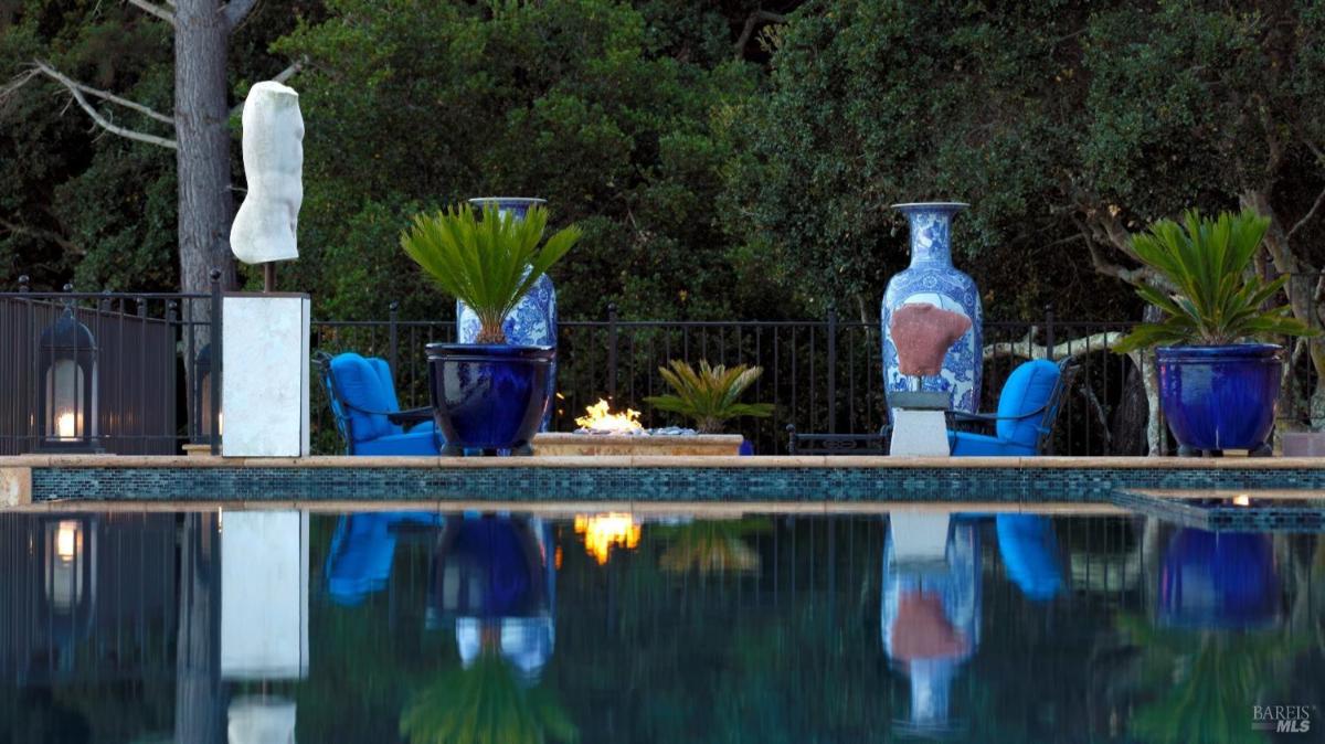 Poolside view of fire pit area with blue ceramic planters, a stone sculpture, and reflections on the water.