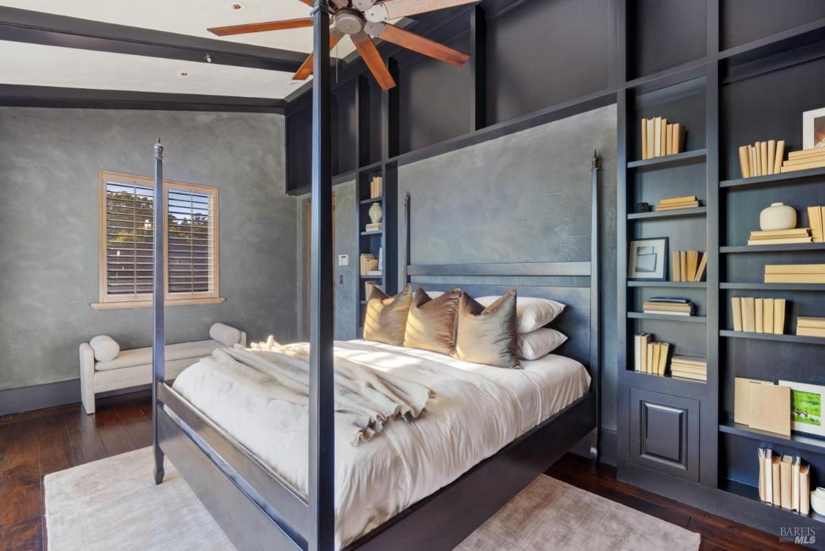 A bedroom featuring a four-poster bed with pillows and a blanket. There is a small seating area and a bookshelf with decorative items.