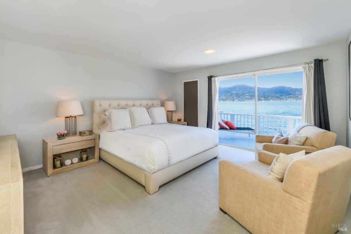 A primary bedroom with a large bed, sitting area, and sliding doors opening to a deck overlooking the water.