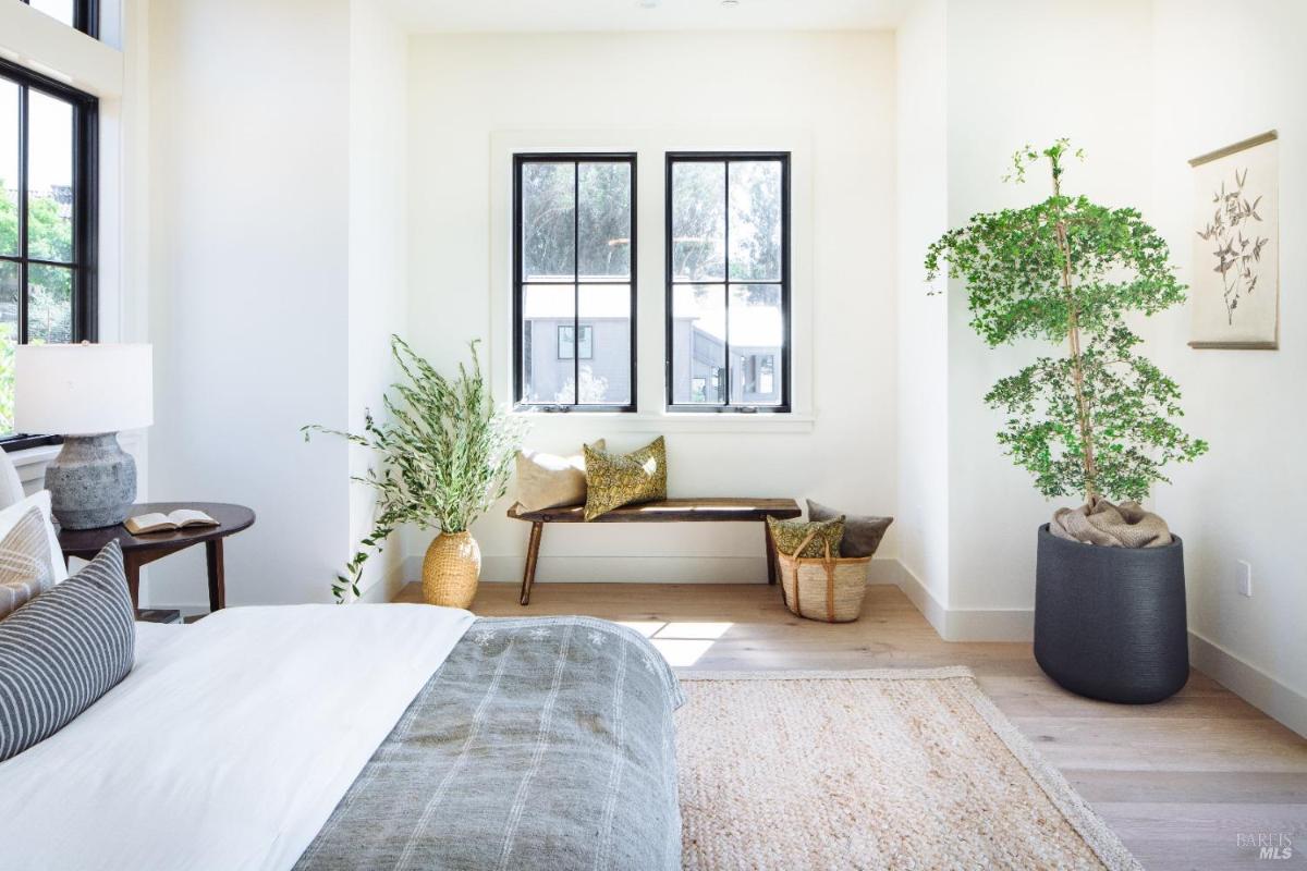 A bedroom with a bed, a bench, plants, and large windows.